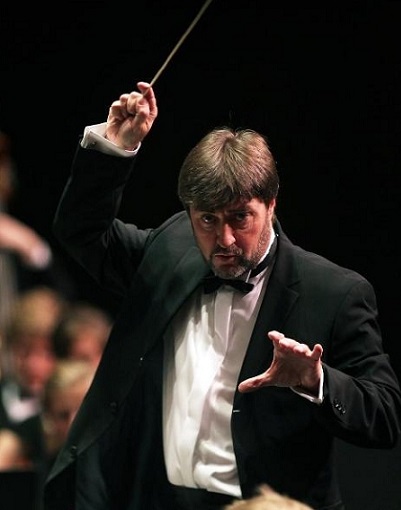 Foto von einem Mann im Frack der einen Dirigierstab in der Hand hält und ein Orchester dirigiert.