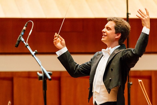 Foto von einem Mann im Frack der einen Dirigierstab in der Hand hält und ein Orchester dirigiert.