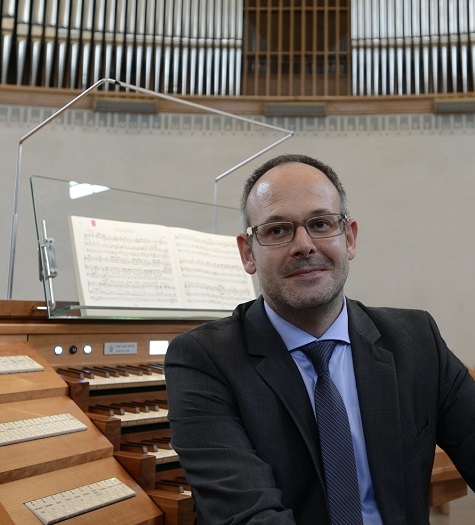 Portraitfoto von einem Mann in Anzug und Krawatte sitzend vor einer Orgel