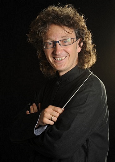 Portraitfoto von einem Mann mit lockigen Haaren, einer Brille. Er hält einen Dirigierstab in der Hand.