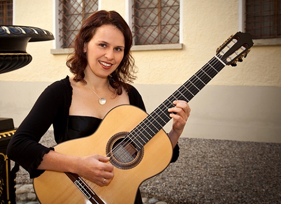 Portraitfoto von Ulrike Schuh, sitzend vor einem Brunnen mit einer Gitarre in der Hand.